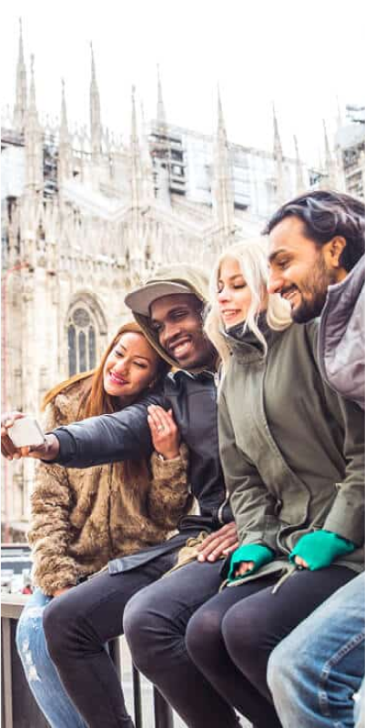 Four people sitting on a ledge, smiling and taking a selfie in front of a large, ornate building