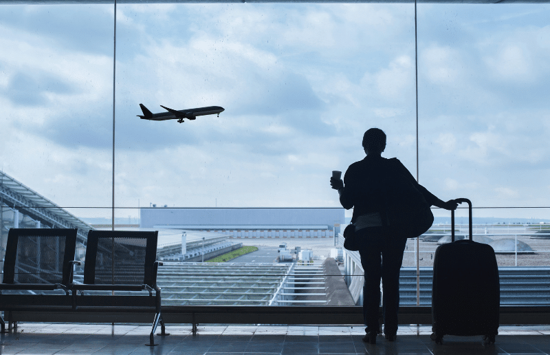 Woman stands facing windows holding a cup of coffee. She is watching a plane fly by in an airport.