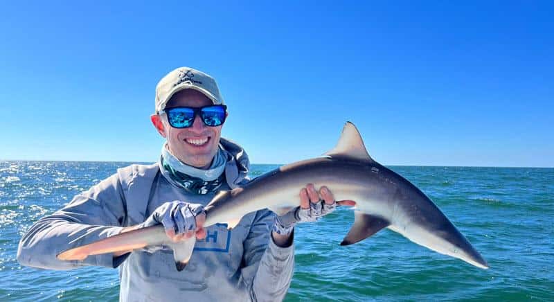 Jason Lenard wearing sun-protective fishing gear and polarized sunglasses smiling while holding a small shark horizontally in front of him on a boat, with calm ocean water and a clear blue sky in the background.